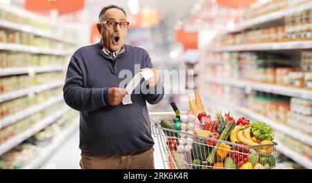 Schockierter reifer Mann mit einem Einkaufswagen, der eine Rechnung im Supermarkt hält Stockfoto