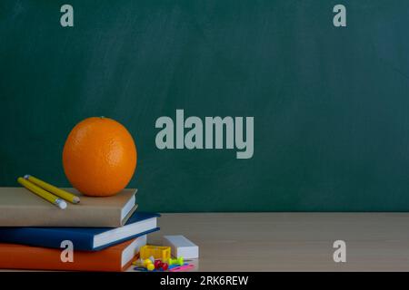 A stack of books, one orange and colorful, school supplies, on a green background of the blackboard with a place for the inscription. Stock Photo