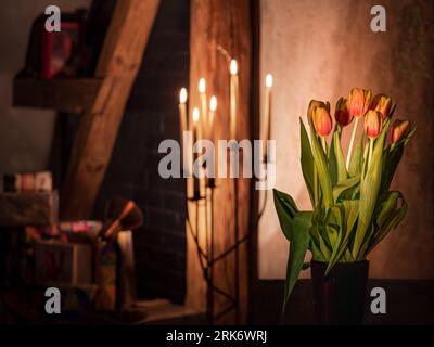 Eine elegante Keramikvase mit rosa und weißen Tulpen steht in einem antiken Raum mit schwach beleuchteten Kerzen und schafft eine geheimnisvolle Atmosphäre Stockfoto