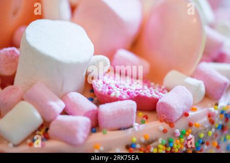 Ein rosafarbener Geburtstagskuchen, dekoriert mit rosa und weißen Marshmallows, Macarons und Donut und bedeckt mit bunten kleinen Streuseln an den Seiten. Stockfoto