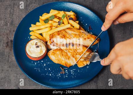 A closeup of a person cutting a chicken steak with a knife, served with fries and sauce Stock Photo