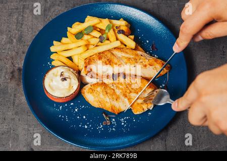 A closeup of a person cutting a chicken steak with a knife, served with fries and sauce Stock Photo