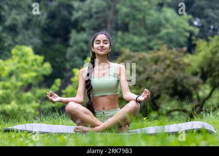 Beautiful young Indian woman doing yoga in the park. Sitting on a mat with closed eyes in the lotus position and meditating. Stock Photo