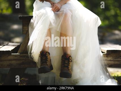 Eine junge Frau, die auf einer Bank sitzt und ein weißes Brautkleid und braune Stiefel trägt Stockfoto