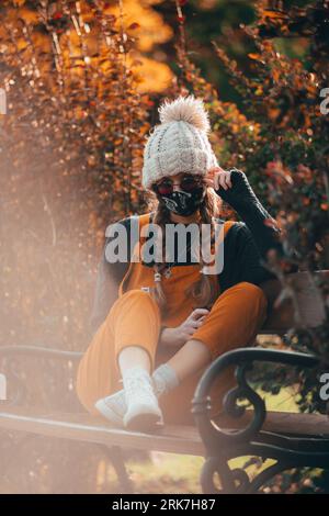 Eine senkrechte blonde Frau mit einer Gesichtsmaske, die im Herbst in einem Park sitzt Stockfoto
