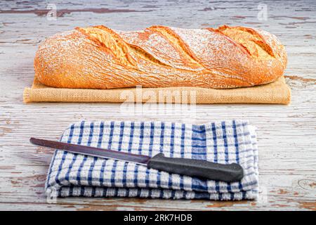 A freshly-baked loaf of bread placed on a sackcloth, with a knife to the side. Stock Photo