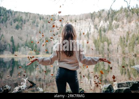 A young woman wearing tight jeans and a sweater, standing in front of a lake, throwing leaves. Stockfoto