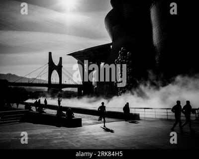 A black and white photograph of a bridge with foggy air emanating from it, situated in front of a landscape of trees and hills Stock Photo