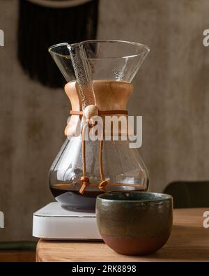 A glass coffee pot sits atop a dark brown wooden table, surrounded by a warm yellow light Stock Photo