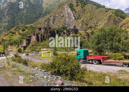 Das befestigte Dorf Shatili (georgisch შატილი) erhebt sich mit seinen Schiefertürmen auf einem felsigen Hang über einer Straße in der Munizipalität Duscheti, Region Mtskheta-Mtianeti, Georgien. Ein grüner LKW mit einem roten Flachbett und einem Anhänger steht im Vordergrund rechts. Ein großer gelber Rahmen steht auf einem Schotterplatz in der Mitte. Straßenschilder stehen am Straßenrand links. Grüne Berge mit felsigen Flanken erheben sich hinter dem Dorf. Die Straße über den Datvisjvari-Pass (wörtliche Übersetzung des georgischen Namens: Bären-Kreuz) ist von Oktober bis Mai meist wegen Schnee gesperrt. Das Dorf liegt auf einer Höhe von etwa Stockfoto