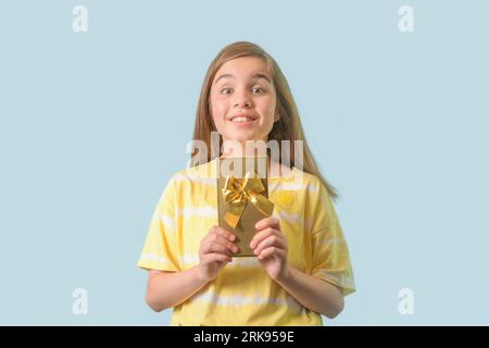 Fröhliches Mädchen im Teenageralter - hält ein Geschenk in einem vergoldeten Paket mit einer Schleife in den Händen. Hellblauer Hintergrund Stockfoto