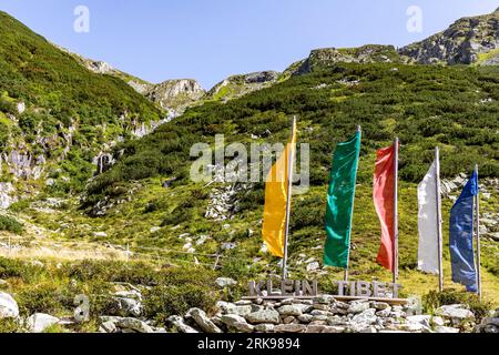 Brandberg, Österreich. August 2023. Fahnen in den Farben tibetischer Gebetsfahnen stehen auf einem Wanderweg am Zillergrund oberhalb der hölzernen Inschrift „KLEIN TIBET“. Der Weg führt zu einer kleinen bewirtschafteten Almweide am Zufluss des Ziller, der dem österreichischen Zillertal in Tirol seinen Namen gibt, in den Stausee. Die Idee für 'Klein Tibet', eigentlich 'Hohenaualm' kam dem Besitzer nach einem Aufenthalt in Tibet wegen der angeblichen Ähnlichkeit der Landschaft. Quelle: Frank Hammerschmidt//Frank Hammerschmidt/dpa/Alamy Live News Stockfoto