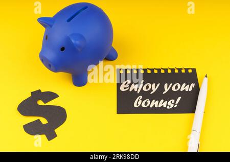 Business concept. On a yellow background there is a piggy bank, next to it is a dollar symbol and a leaf from a notepad with the inscription - Enjoy y Stock Photo