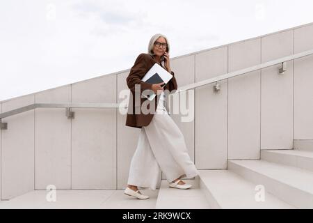 Selbstbewusste reife alte Dame mit grauen Haaren, die eine Brille trägt, die eine braune Jacke trägt, geht zum Geschäftstreffen auf der Treppe Stockfoto