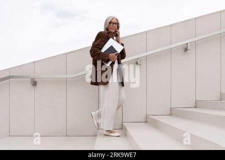 Eine grauhaarige Frau mittleren Alters mit bob-Frisur in Jacke und Hose spaziert vor dem Hintergrund eines Business Centers durch die Stadt Stockfoto