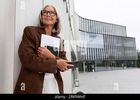Schöne erfolgreiche reife Geschäftsfrau mit grauen Haaren und Dokumenten in den Händen steht vor dem Hintergrund eines Business Centers Stockfoto