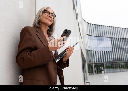 Eine gepflegte Frau mittleren Alters in Gläsern, die in einer braunen Jacke gekleidet ist, steht an der Wand eines Business Centers Stockfoto