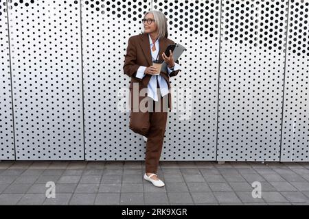 Eine Geschäftsfrau reifer Jahre, die in einer eleganten braunen Jacke, Hose und einem Hemd gekleidet ist, spaziert durch die Straße der Stadt Stockfoto