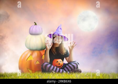 Kleines Mädchen, das an Halloween auf einer riesigen Kürbislaterne sitzt. Riesiger Vollmond im Hintergrund. Das Hexenkostüm für Kinder ist ein Trick oder ein Genuss. Kinder haben Fu Stockfoto