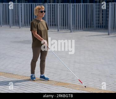 Eine ältere blinde Frau läuft mit einem Stock auf einem taktilen Fliesen. Stockfoto