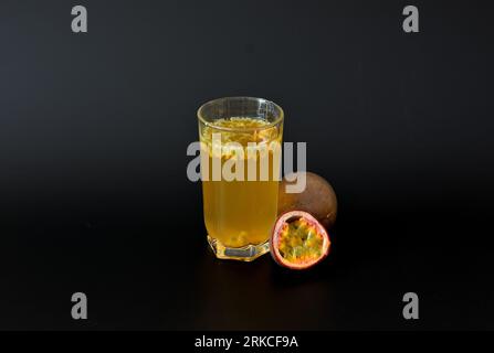 A glass of tropical fruit juice with seeds on a black background, next to pieces of ripe passion fruit. Close-up. Stock Photo