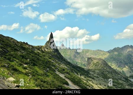 A gentle slope covered with moss and grass and two pointed peaks of a steep cliff on a sunny summer day. Parabola Rock, Ergaki Nature Park, Krasnoyars Stock Photo