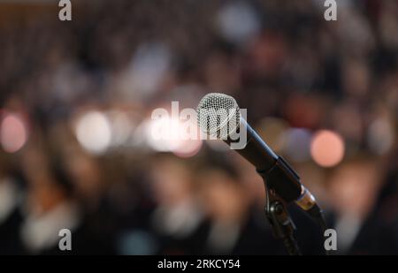 Close up of a microphone on stage with a burred bokeh audience or crowd. Public speaking performing nervousness or anxiety theme Stock Photo