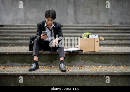 Stressed and depressed young Asian businessman or male office worker sits on a staircase with a box of his personal stuff after getting fired or losin Stock Photo