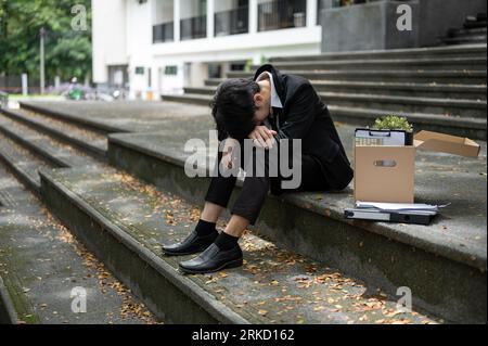 A depressed and stressed young Asian businessman sits on the stairs in front of the building with a box of his personal stuff after getting fired. Job Stock Photo