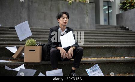 Ein depressiver und trauriger junger asiatischer Geschäftsmann sitzt auf der Treppe vor dem Firmenbüro mit einer Schachtel seiner persönlichen Sachen, nachdem er seinen Job verloren hat Stockfoto