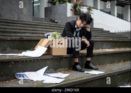 Ein depressiver und trauriger junger asiatischer Geschäftsmann sitzt auf der Treppe vor dem Firmenbüro mit einer Schachtel seiner persönlichen Sachen, nachdem er seinen Job verloren hat. Stockfoto