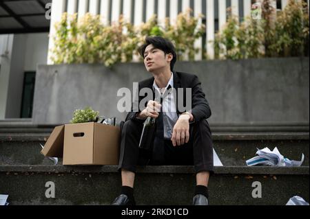 Ein betrunkener, depressiver und gestresster asiatischer Geschäftsmann, der auf Treppen vor dem Firmengebäude mit einer Schachtel seiner persönlichen Sachen sitzt Stockfoto