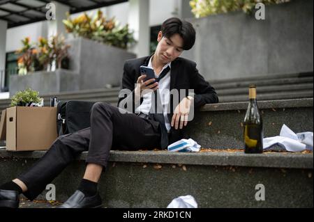 A drunk and hopeless Asian unemployed businessman is typing on his phone while sitting on the stairs in front of the building with a box of his person Stock Photo