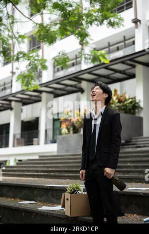 A drunk and stressed Asian businessman is screaming with hopelessness, feeling sad and hopeless after losing his job, standing on the stairs in front Stock Photo