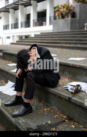 Ein betrunkener, depressiver und hoffnungsloser asiatischer Geschäftsmann sitzt auf der Treppe vor dem Firmengebäude, nachdem er seinen Job verloren oder gefeuert wurde. A alc Stockfoto