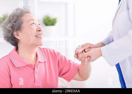 physician leaning forward to smiling elderly lady patient holding her hand in palms. Woman caretaker in white coat supporting encouraging old person Stock Photo