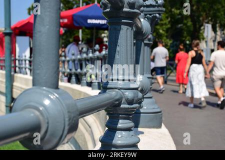 Graues Gusseisengeländer entlang des öffentlichen Asphaltgeheges. Verschwommene Fußgänger, die vorbeifahren. Steinsockel. Sommerszene auf der beliebten städtischen Straße in Budapest Stockfoto