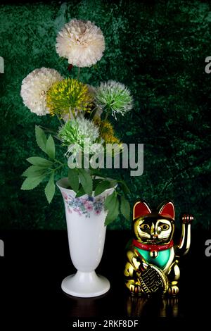 Synthetische Blumen in einer glasierten, verzierten Blumenvase mit goldenem maneki-Neko locken die Glückskatze auf eine hölzerne Tischplatte Stockfoto