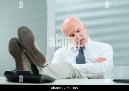 Due to neglect or exhaustion, a senior businessman heavily sleeps in his office, drooling. His feet are even on the desk. Perhaps he's been there so l Stock Photo