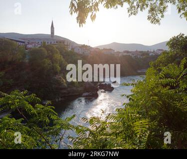 Blick zwischen üppigen Bäumen am Fluss Neretva in Mostar, mit einem Dorf und einer Moschee am Flussufer, Bosnien und Herzegowina, 22. August 2023. Stockfoto