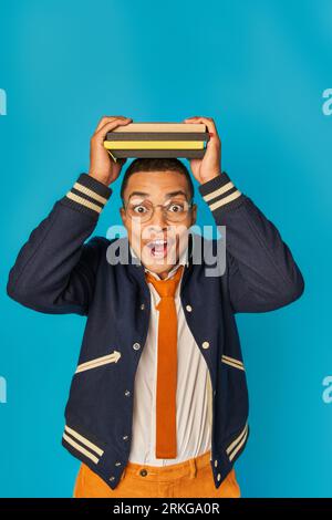 astonished african american student in trendy jacket and eyeglasses with notebooks on head on blue Stockfoto