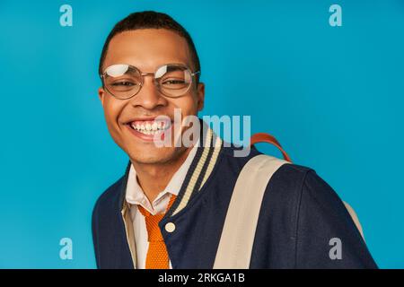 pleased african american student in eyeglasses and trendy jacket smiling at camera on blue, portrait Stockfoto