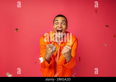 overjoyed african american man in orange shirt shouting and showing win gesture near confetti on red Stockfoto