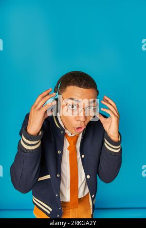 astonished african american student in headphones, with open mouth looking at camera on blue Stockfoto