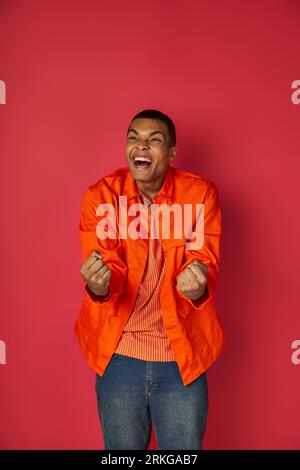 delighted, excited african american shouting and showing win gesture, orange shirt, red background Stockfoto