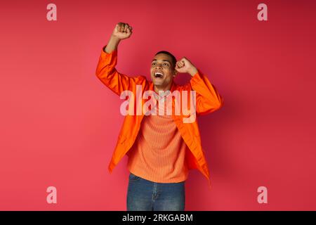 excited african american guy in orange shirt showing success gesture and shouting on red background Stockfoto