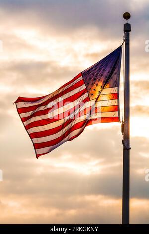 Stars and stripes American flag flying on a flag pole, backlit by sunlight against a sky Stock Photo