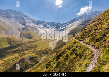 Ein schmaler Pfad verläuft entlang eines steilen, grünen Hangs auf dem Abstieg vom Atsunta-Pass auf der Khevsur-Seite nahe dem Dorf Andaki in der Gemeinde Duscheti, Region Mzcheta-Mtianeti, Georgien. Ein Wanderer mit Kappe ist auf dem Weg entlang des Pfades. Unten öffnet sich ein breites Tal mit einem hellen Kiesbett. Graue Felspitzen mit Schneeflecken erheben sich dahinter. Links fließt ein Bach durch eine Schlucht. Zwei weiße Wolken hängen im blauen Himmel über den Bergen. Die Trekkingtour mit Packpferden hat fast 120 Kilometer und mehr als 5.000 Meter Auf- und Abstieg zurückgelegt. Khewsureti (georgisch ხევსურეთი) ist eine historische Region Stockfoto