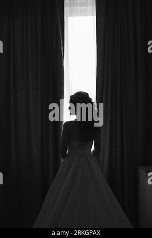 A full-length shot of a bride standing in profile, admiring the view from a tall window in her wedding dress Stock Photo