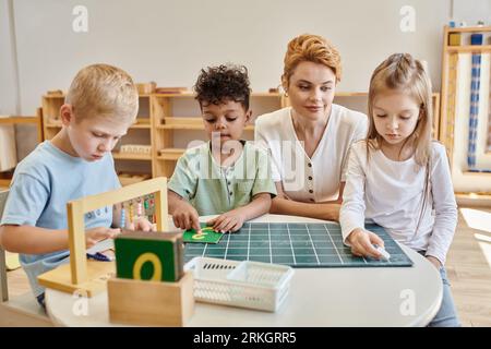 montessori Schulkonzept, multikulturelle Kinder, Mädchen schreiben auf Tafel in der Nähe von Lehrer und Jungen Stockfoto